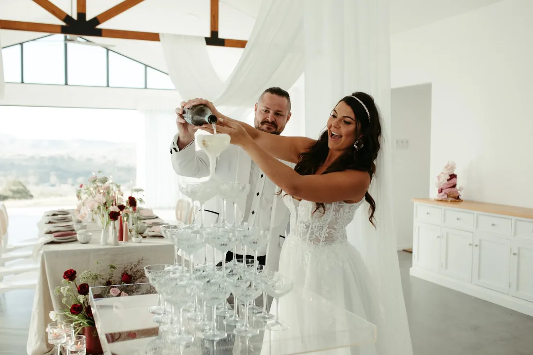 Bride and groom laughing while pouring champagne into a tower of glasses inside the reception room