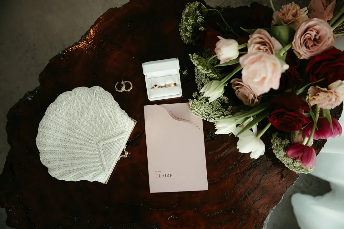 Flatlay of a beaded clutch, gold rings, a Claire place card and a bouquet of burgundy and blush blooms