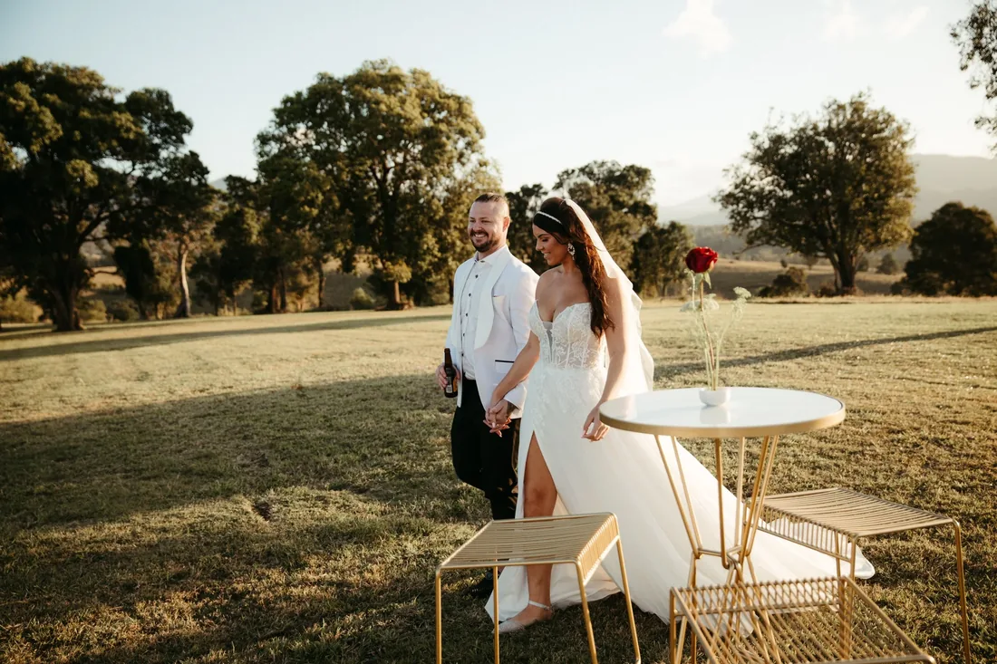 Bride and groom standing beside a gold cocktail table styled with a single red rose in golden-hour light