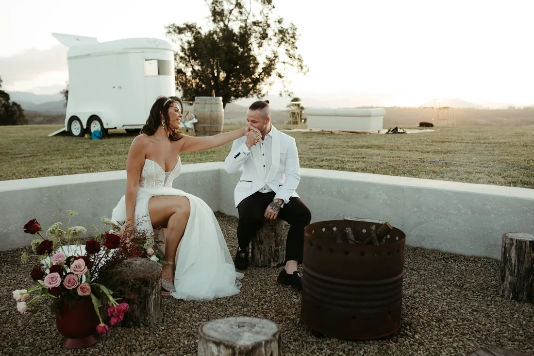 Bride resting her hand on the groom's chin beside a horse float and fire pit at dusk