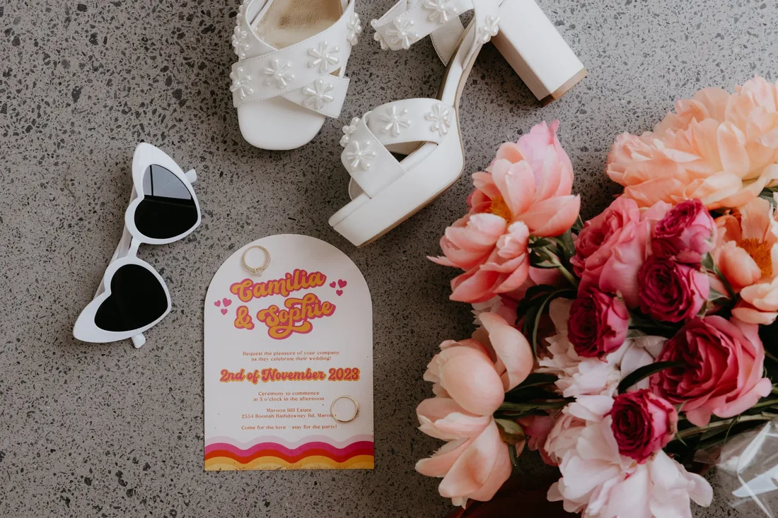 Flatlay of floral platform heels, heart-shaped sunglasses, a retro invitation for Camilla and Sophie and a bouquet of pink and peach blooms