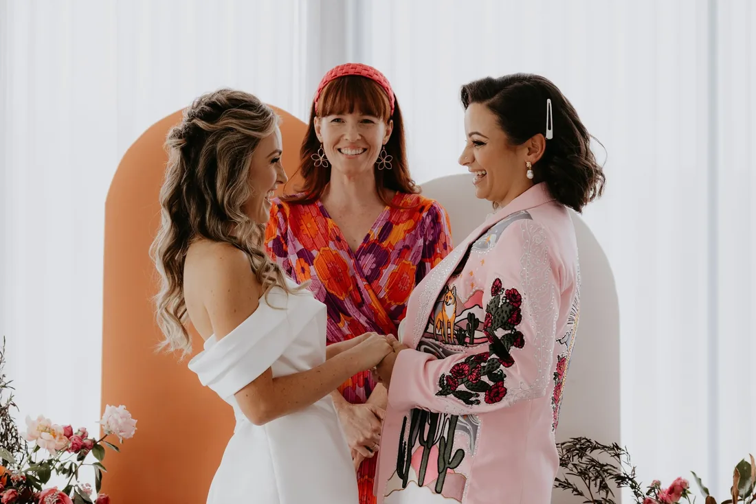 Two brides laughing together with the celebrant during their ceremony, orange arch backdrop