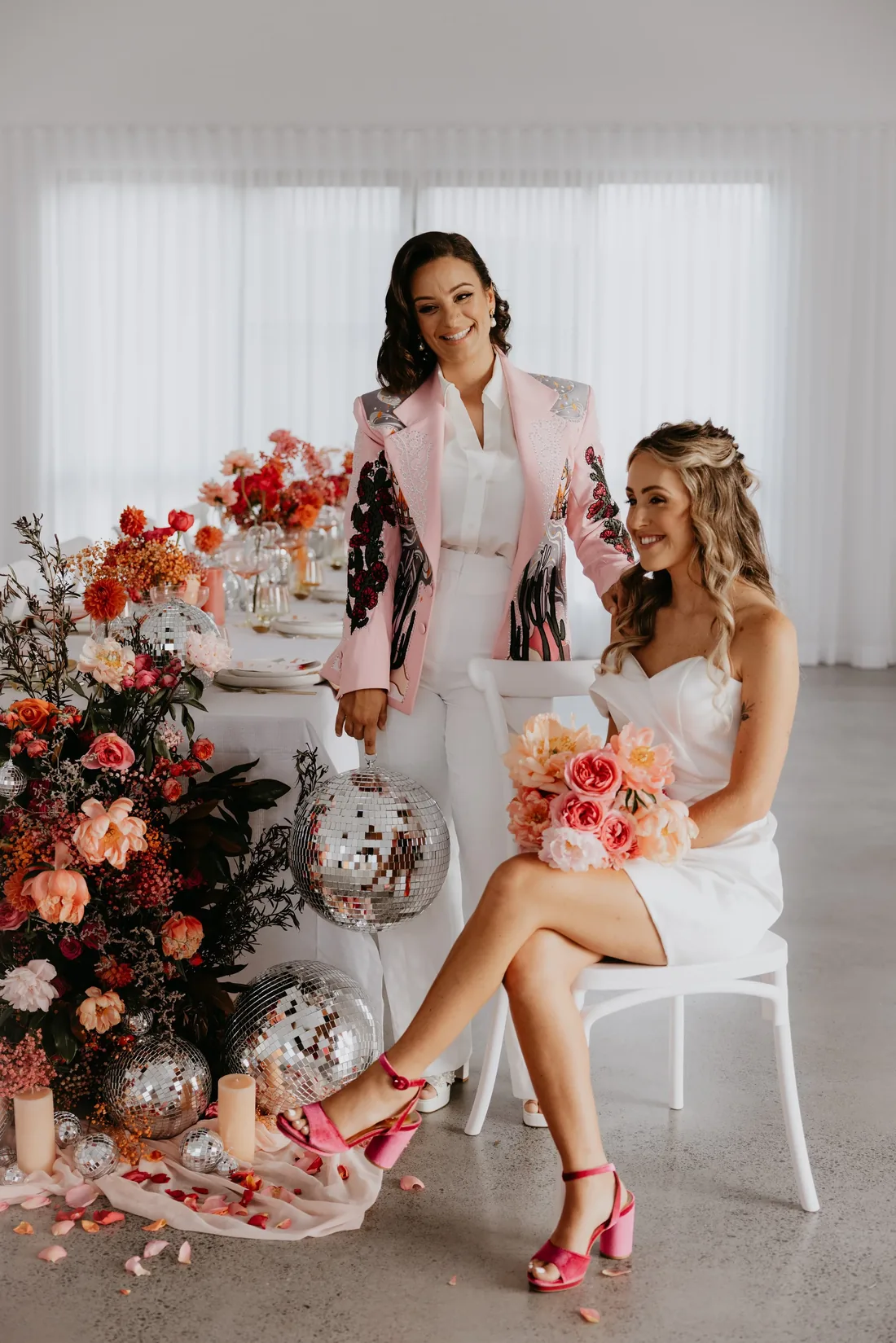 Bride in a pink sequinned blazer helping her partner into her heels beside the styled floral table