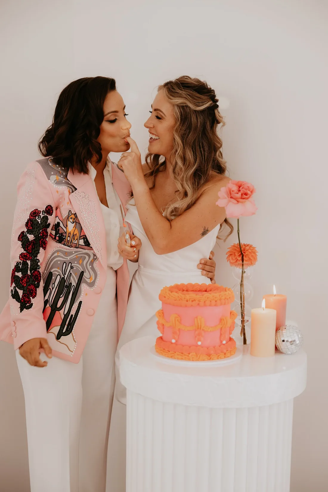 Two brides sharing a playful bite of cake, one in a pink embroidered blazer