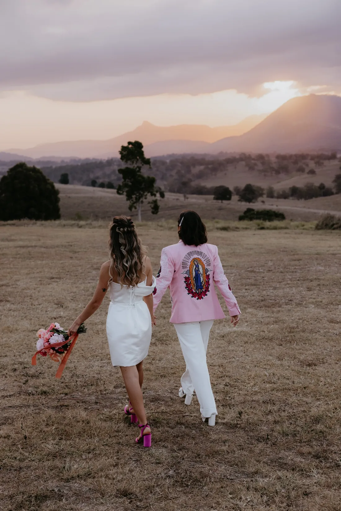 Two brides walking hand in hand across the paddock at sunset, mountain silhouette behind