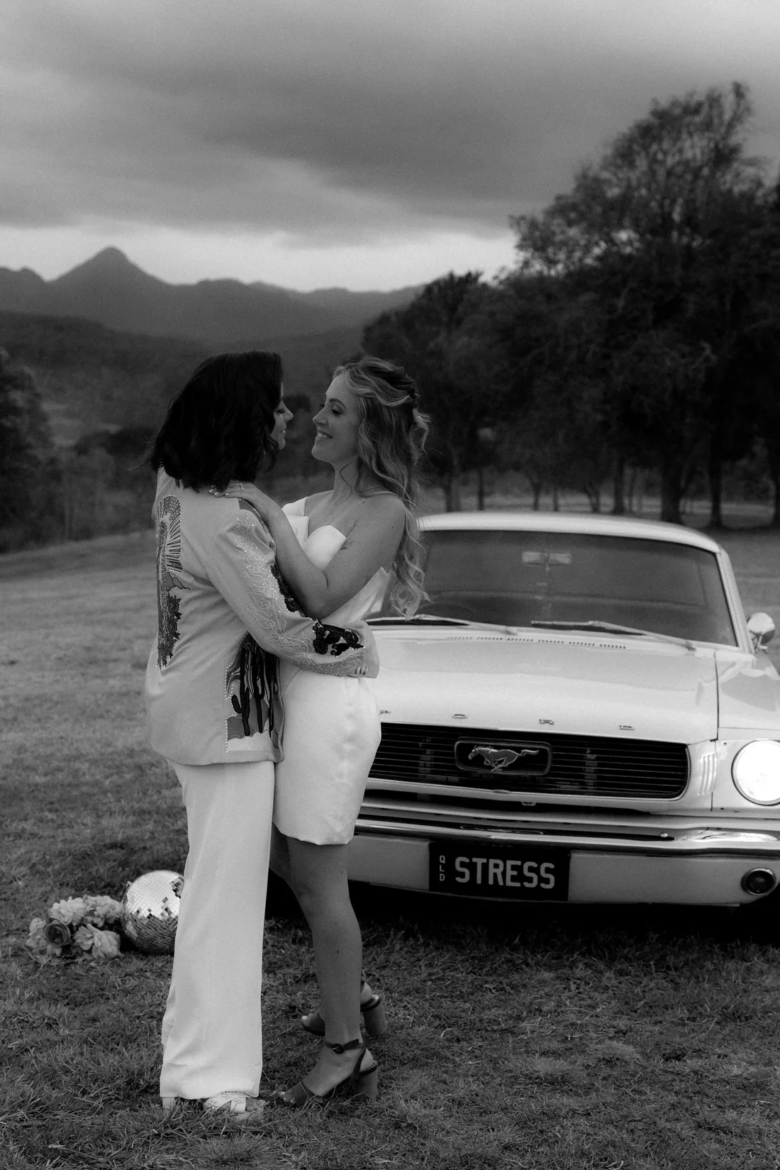 Black and white photo of two brides embracing beside a vintage Mustang with an Old Stress number plate