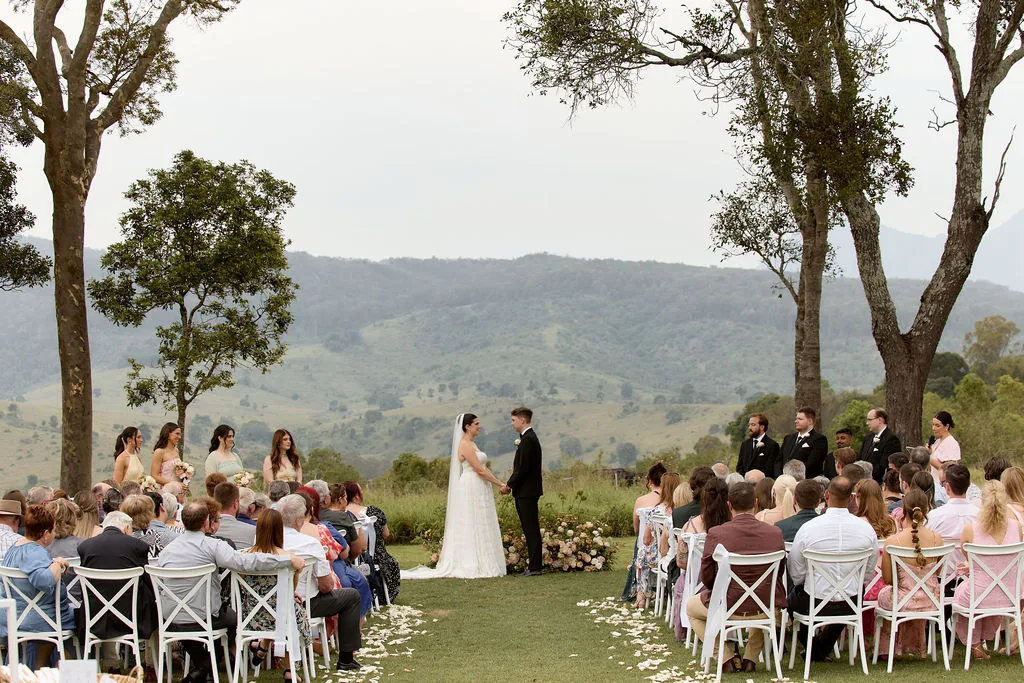 Rebecca and Benjamin holding hands during their vows, bridesmaids and guests either side, mountain views behind