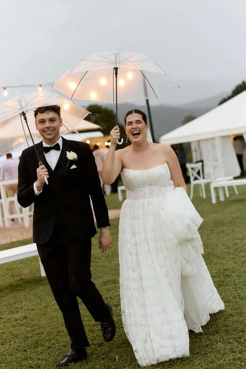 Rebecca and Benjamin laughing under clear umbrellas in the rain, festoon lights behind