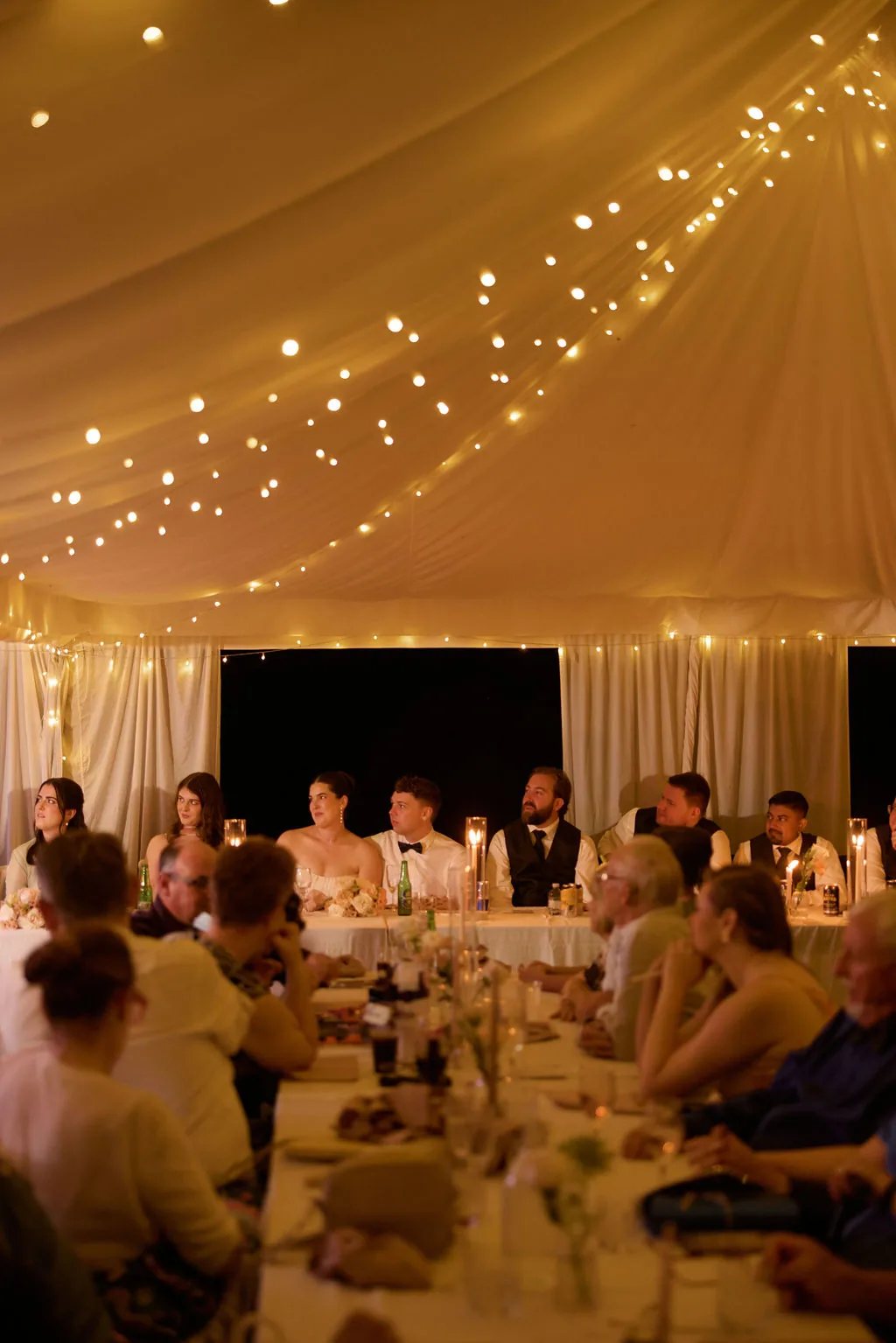 Guests seated at the reception table, laughing during speeches under the marquee's fairy lights