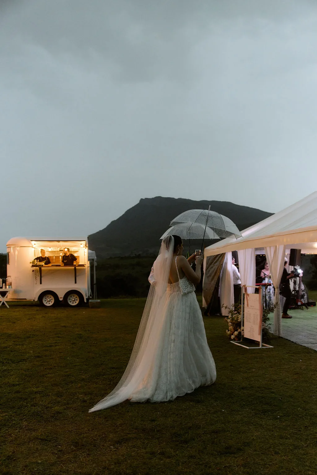 Bride walking under an umbrella past the mobile bar at dusk in the rain