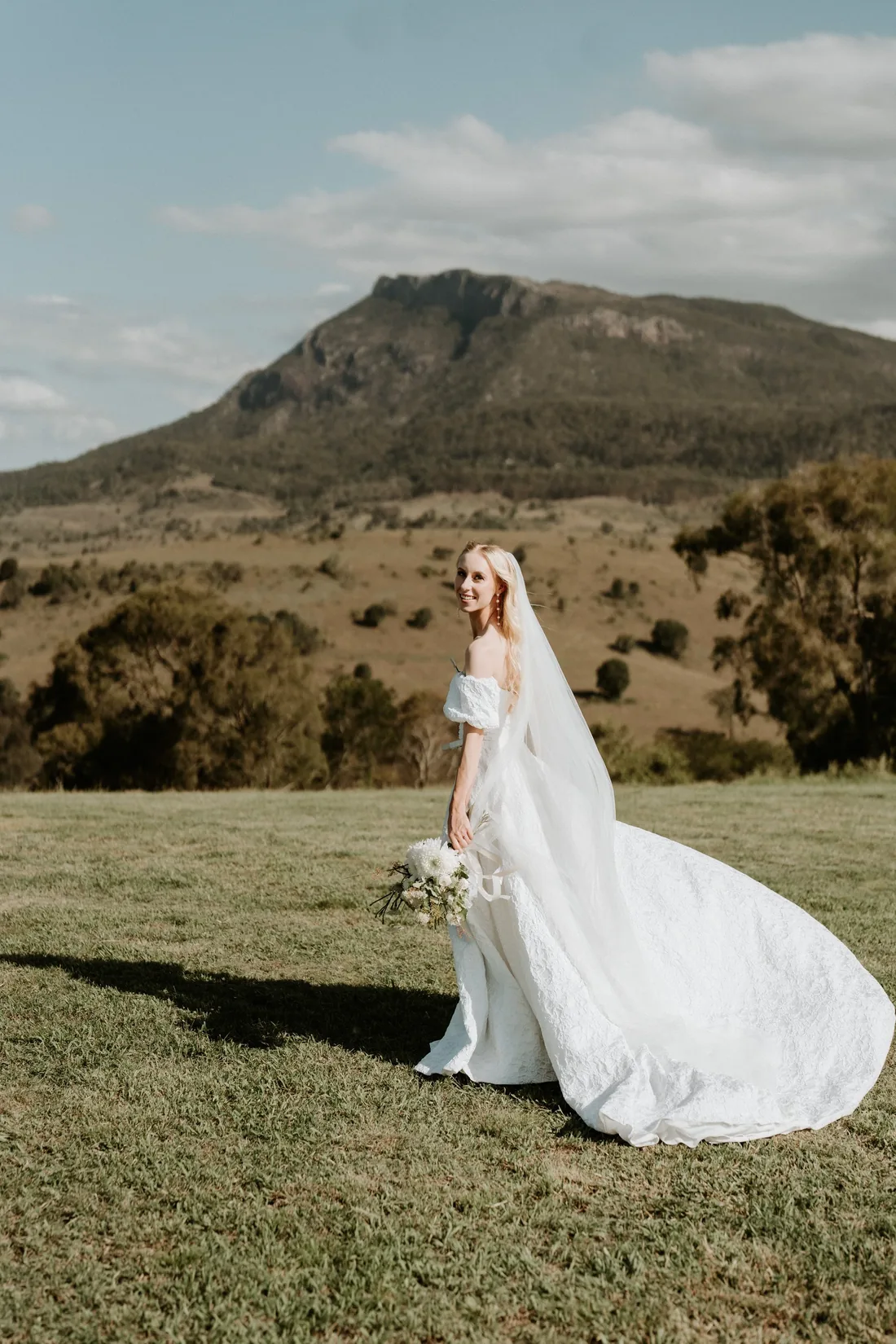Bride walking through the grass with her cathedral veil trailing behind her, mountain backdrop