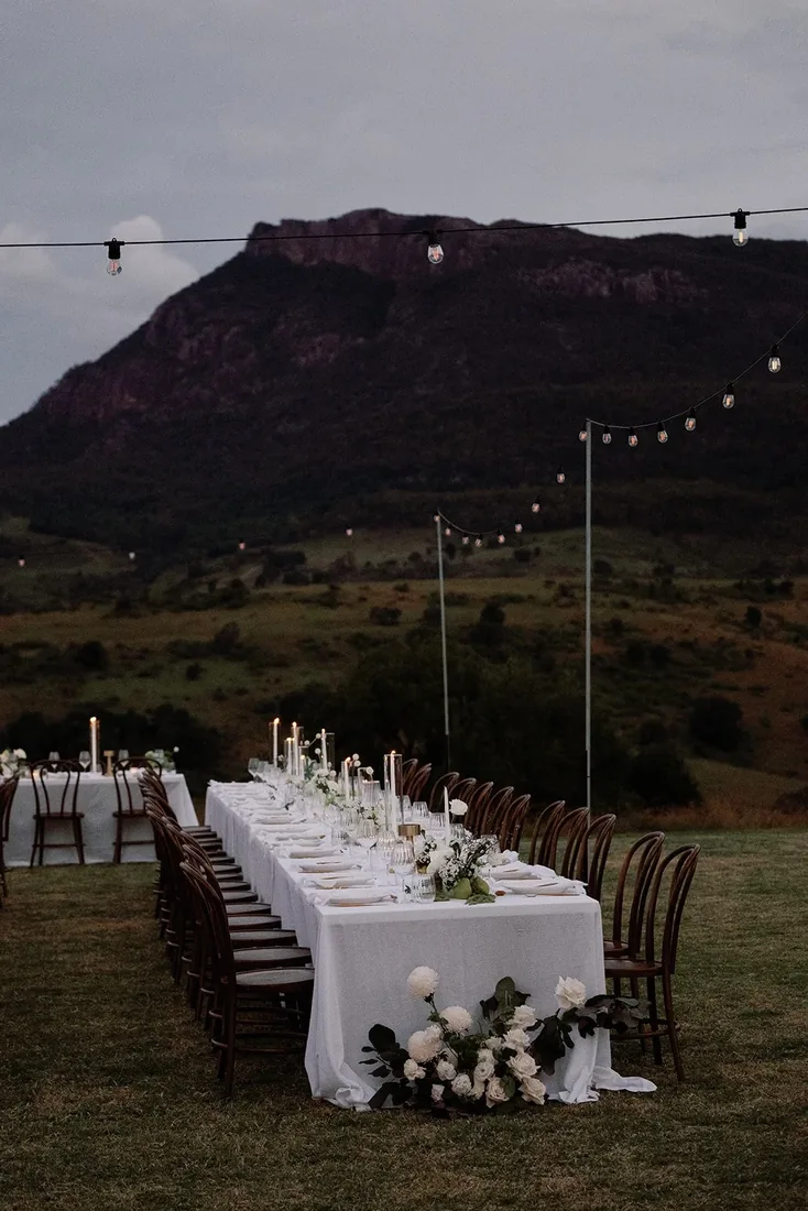 Long reception table set for dinner outdoors at dusk, string lights and a mountain silhouette behind