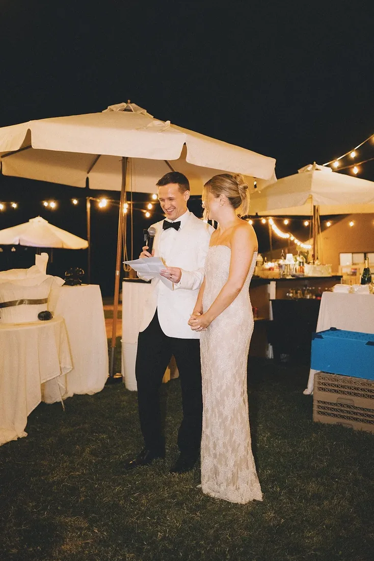 Ryan giving a speech in a white dinner jacket, Samara beside him, market umbrellas and string lights