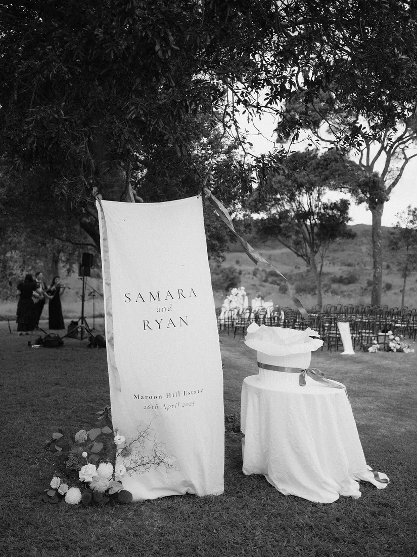 Black and white photo of the Samara and Ryan ceremony sign styled under a tree with florals