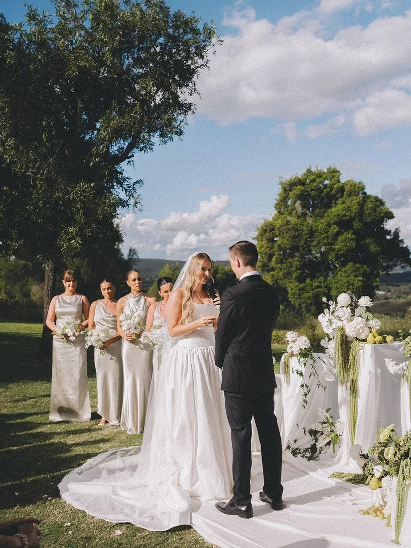 Samara and Ryan exchanging vows at the ceremony, bridesmaids and a floral arbour beside them