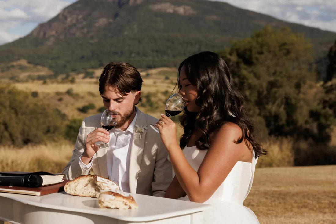 Hamish and Sarah sharing wine and bread during their ceremony, mountain views behind
