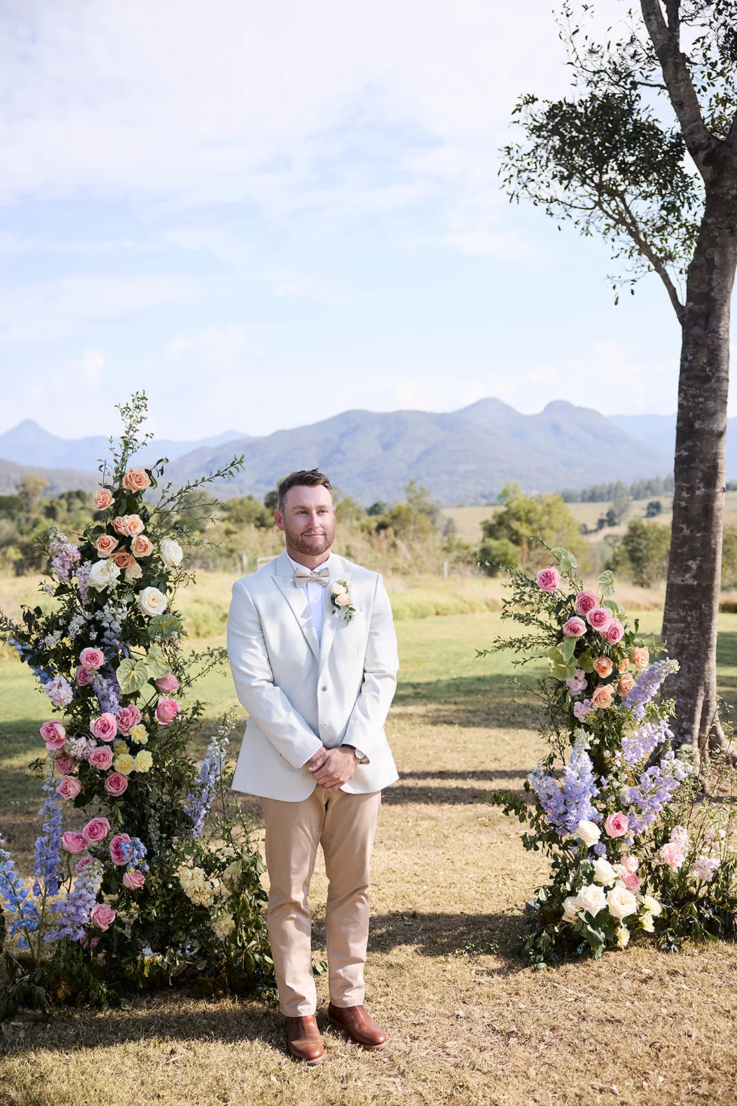 Jayden standing between the two floral ceremony arches in a white blazer, waiting for his bride