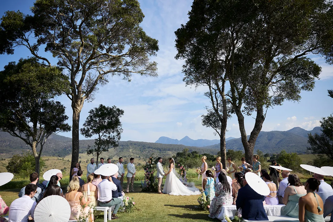 Wide view of the ceremony beneath gum trees, guests seated under white parasols with mountains behind