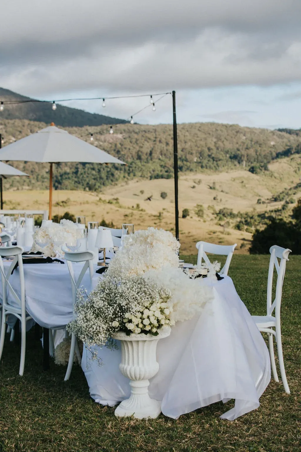 Reception table set beneath market umbrellas with a lush white floral runner, mountain views