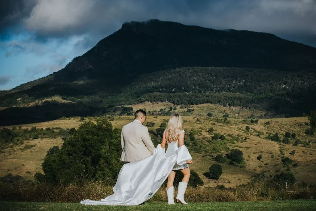 Groom and flower girl looking out over the mountains together, bride's dress trailing behind
