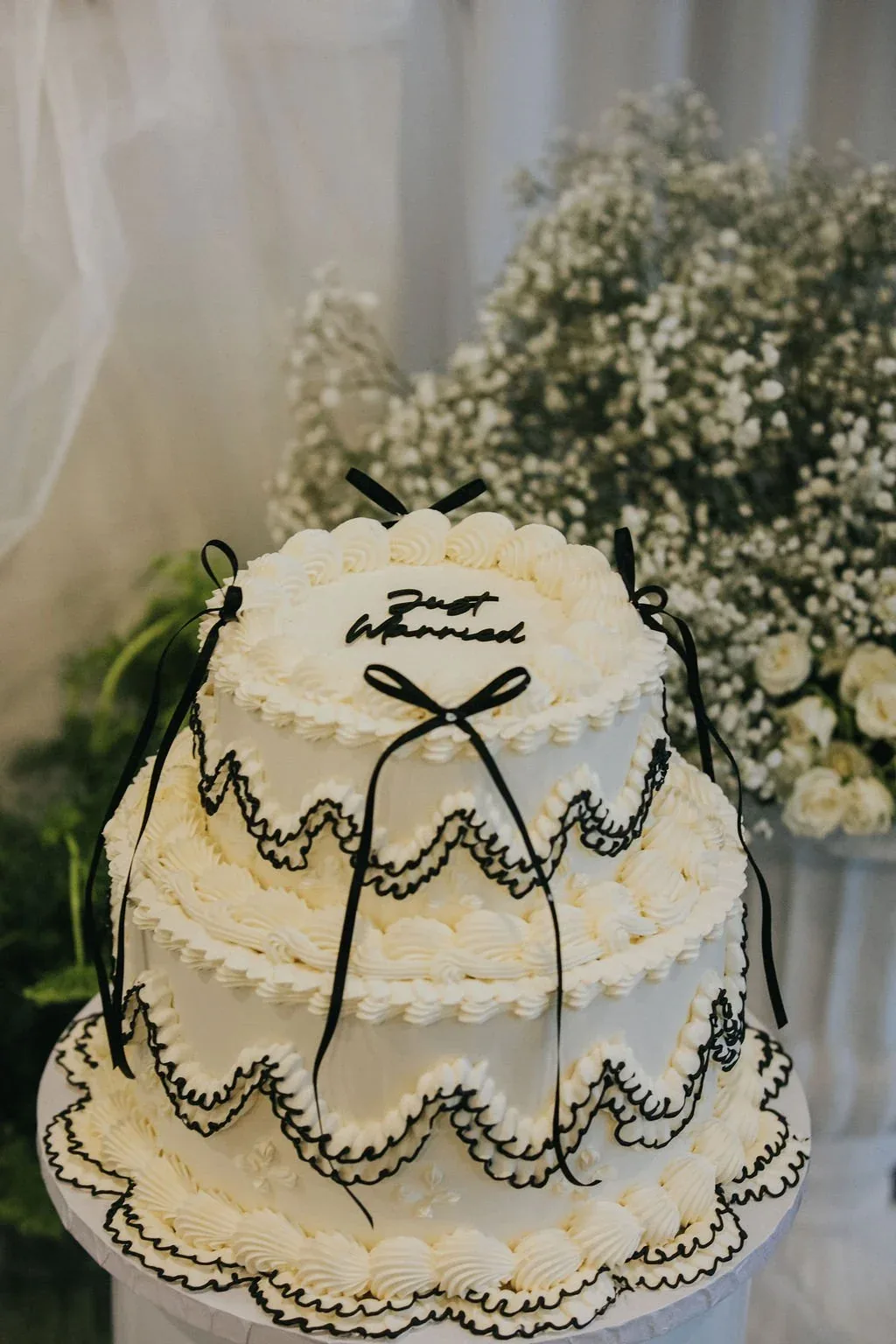 Three-tiered Just Married cake with black ribbon piping and a bow, styled beside white florals