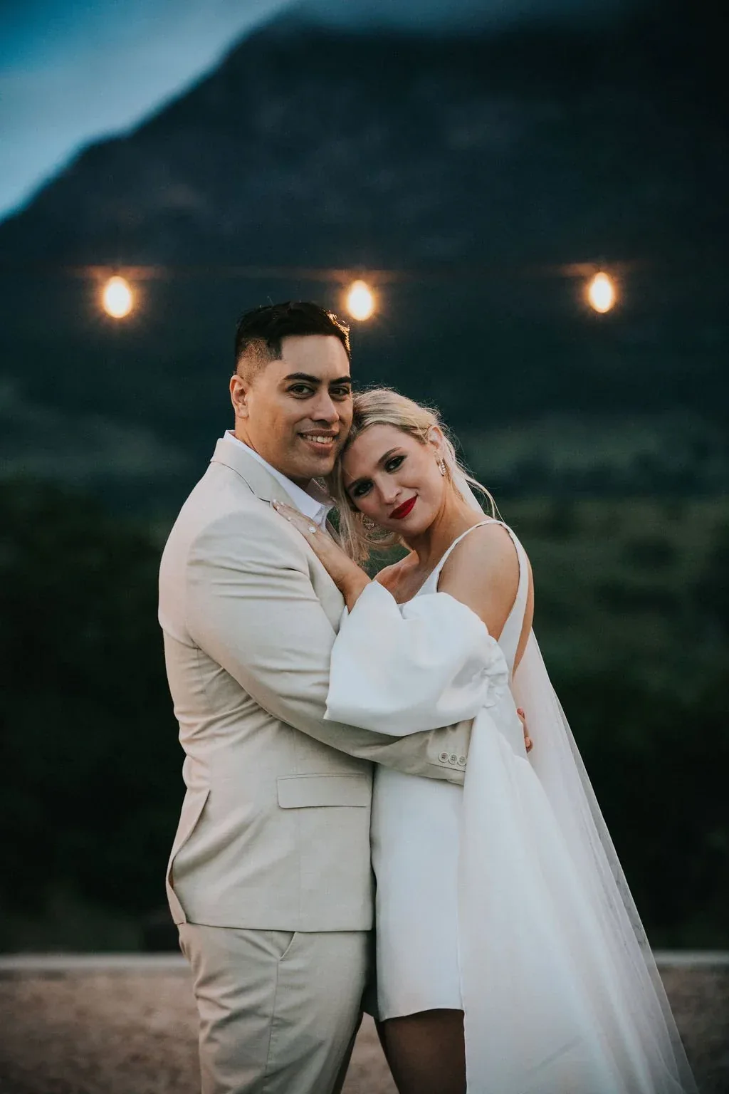 Bride and groom embracing at dusk with string lights and the mountain silhouette behind them