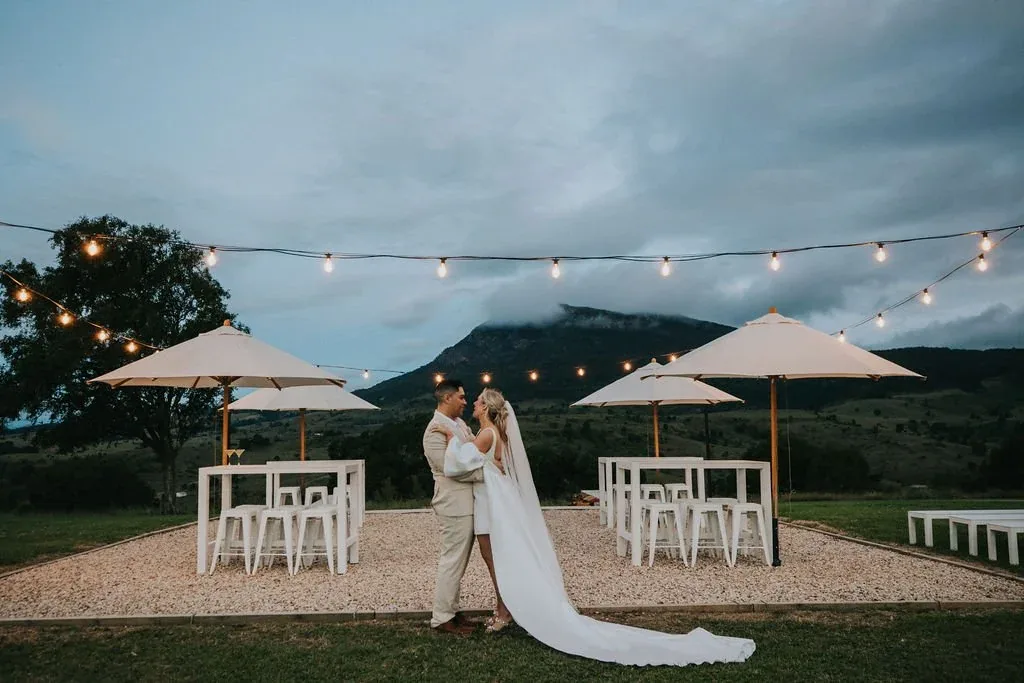 Bride and groom standing together at dusk beneath festoon lights and market umbrellas
