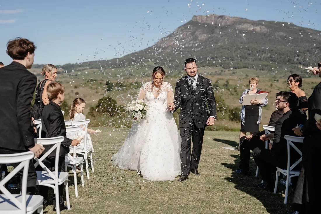 Tylah and Brock walking down the aisle through confetti as guests celebrate, mountain backdrop