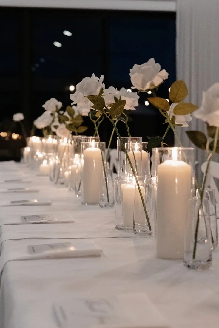 Reception table styled with single white roses in glass vases and lit pillar candles at night