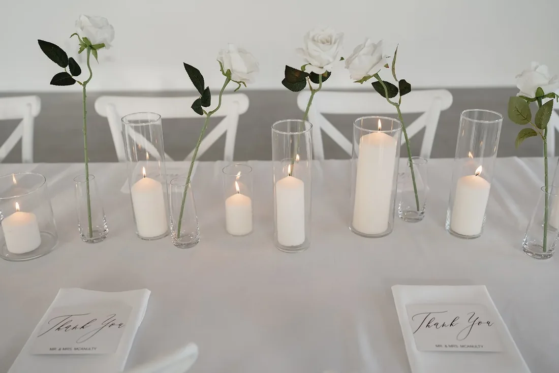 Reception table setting with single white roses in bud vases and lit taper and pillar candles