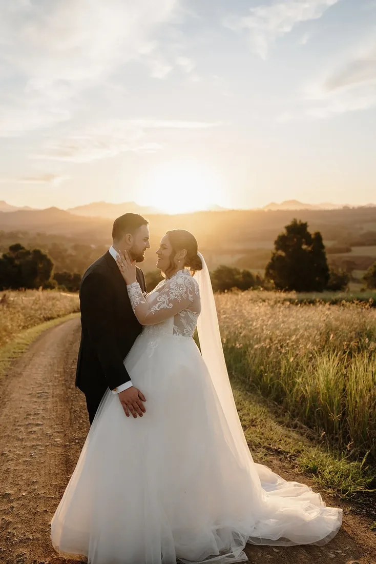 Tylah and Brock embracing on a gravel driveway at sunset, golden light and mountain views