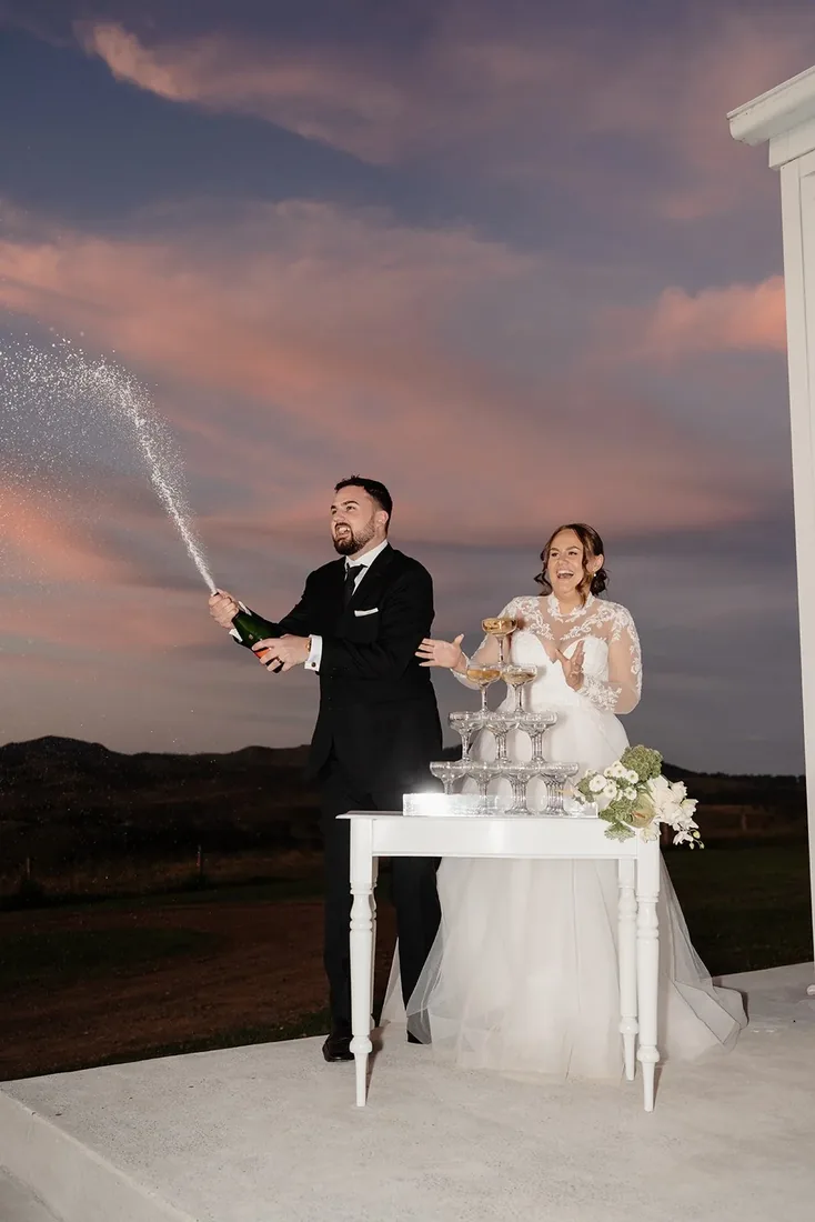 Brock spraying champagne as Tylah laughs beside a tower of glasses at sunset