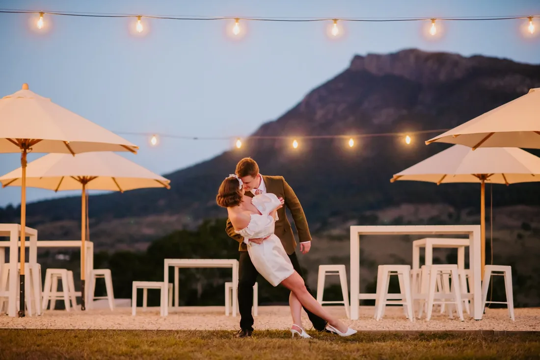 Couple sharing a dip kiss on the lawn at dusk, festoon lights and market umbrellas around them
