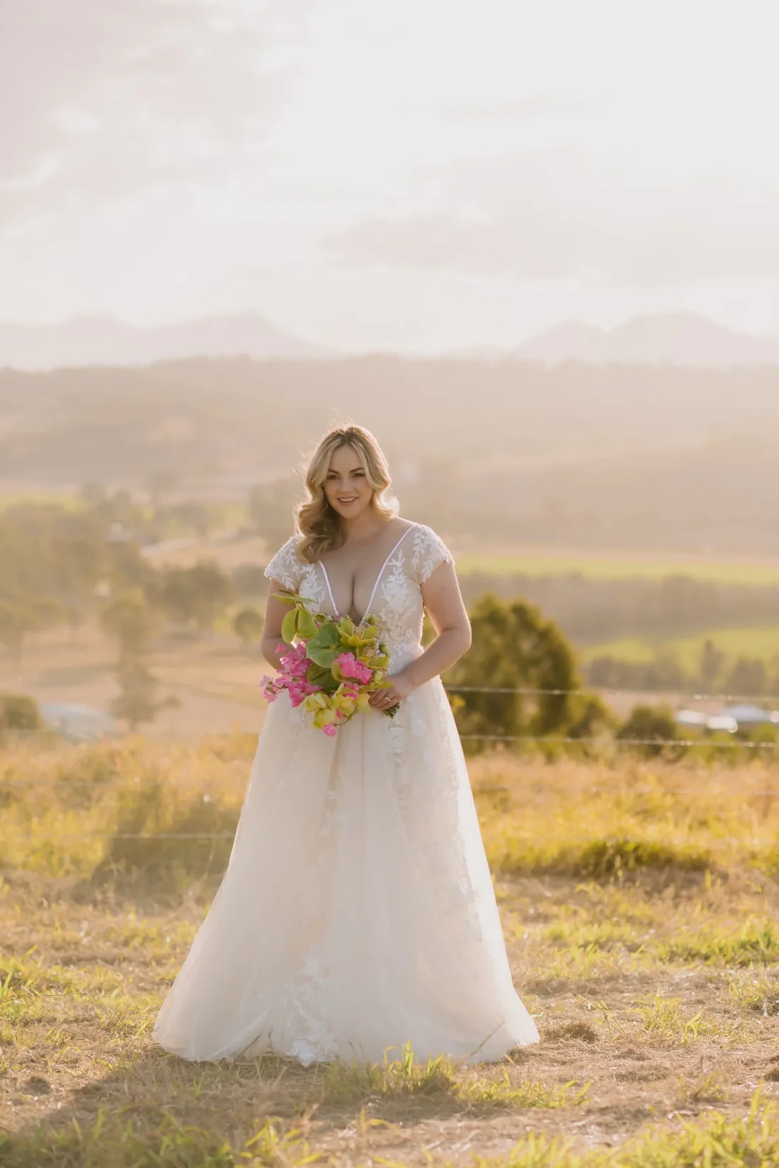 Bride in a lace gown holding a tropical pink and green bouquet, standing in a golden paddock