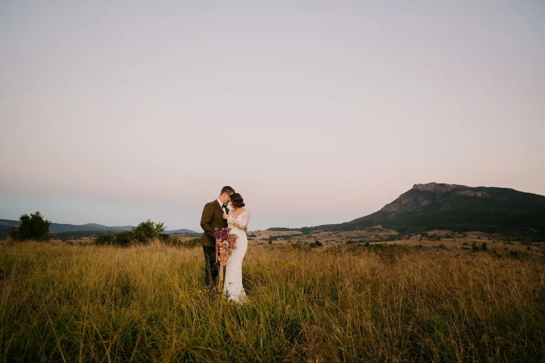 Couple embracing in the long grass at sunset with a mountain silhouette behind them