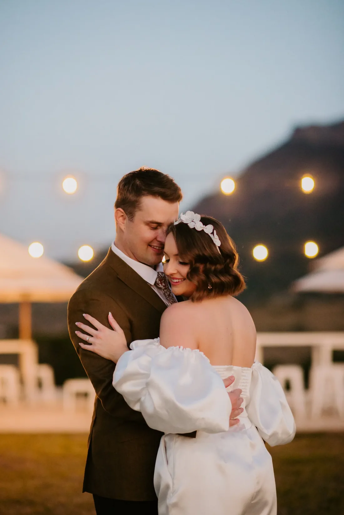 Couple embracing at dusk, string lights and the mountain silhouette glowing behind them