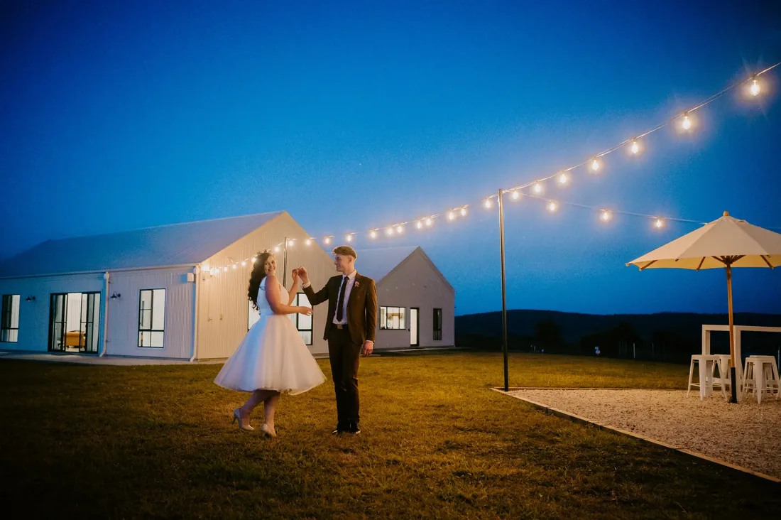Couple dancing together on the lawn at night beneath festoon lights, marquee behind them
