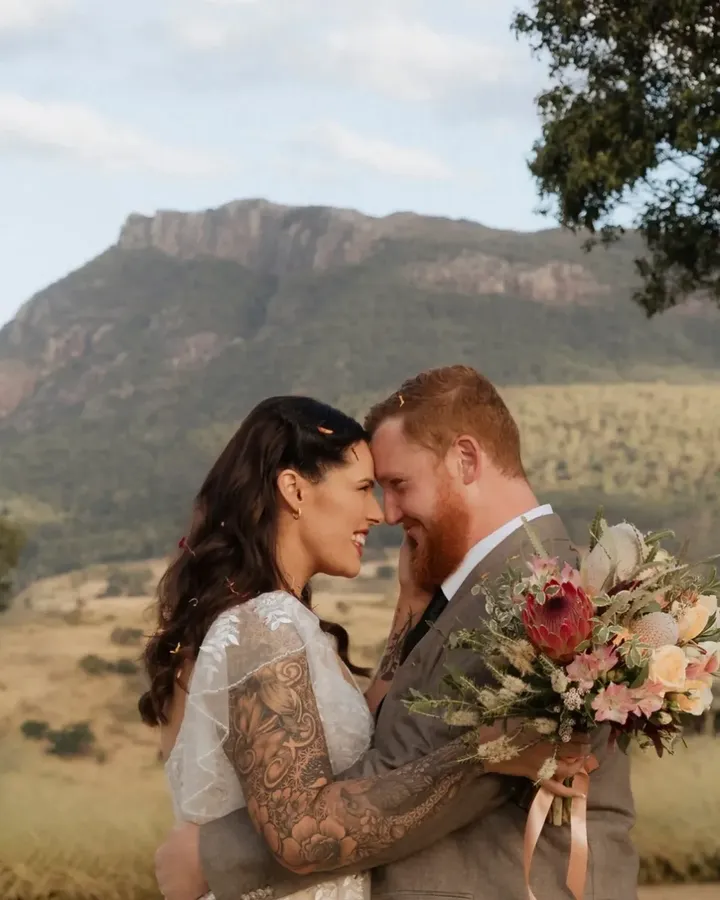 Abby and Liam embracing with a protea bouquet, Mount Maroon behind them